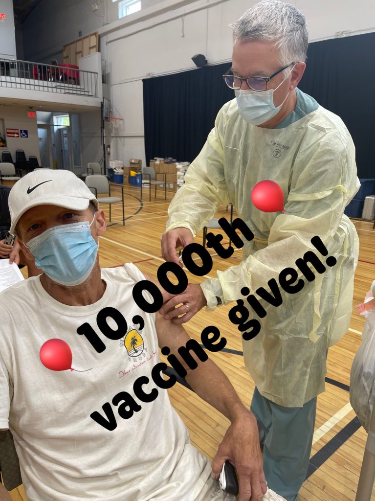 Elliott Lake FHT clinician in PPE administering their 10000th dose of the COVID-19 vaccine to a masked man sitting down