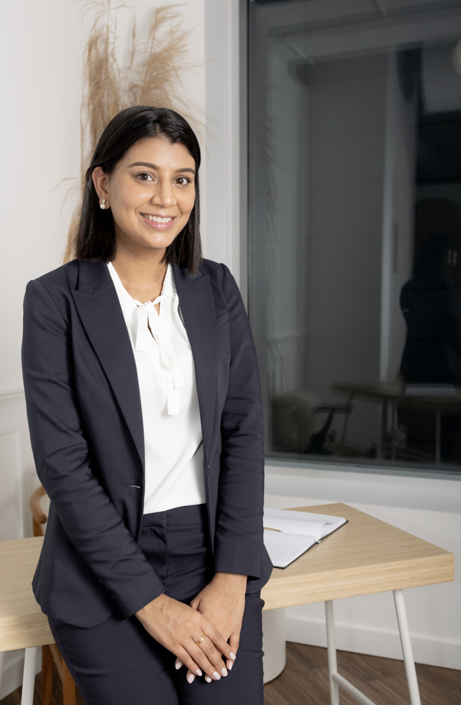 Sandeep perches on edge of desk with her hands in her lap wearing a suit