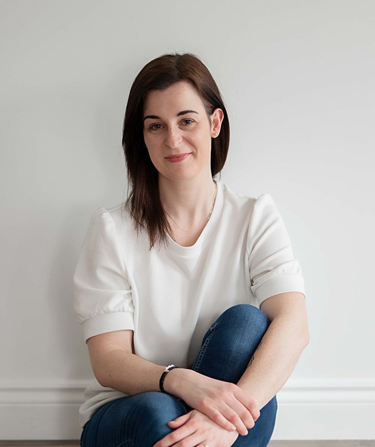 Taryn is wearing a white T-shirt and jeans. She's sitting on the floor with her back against a white wall.