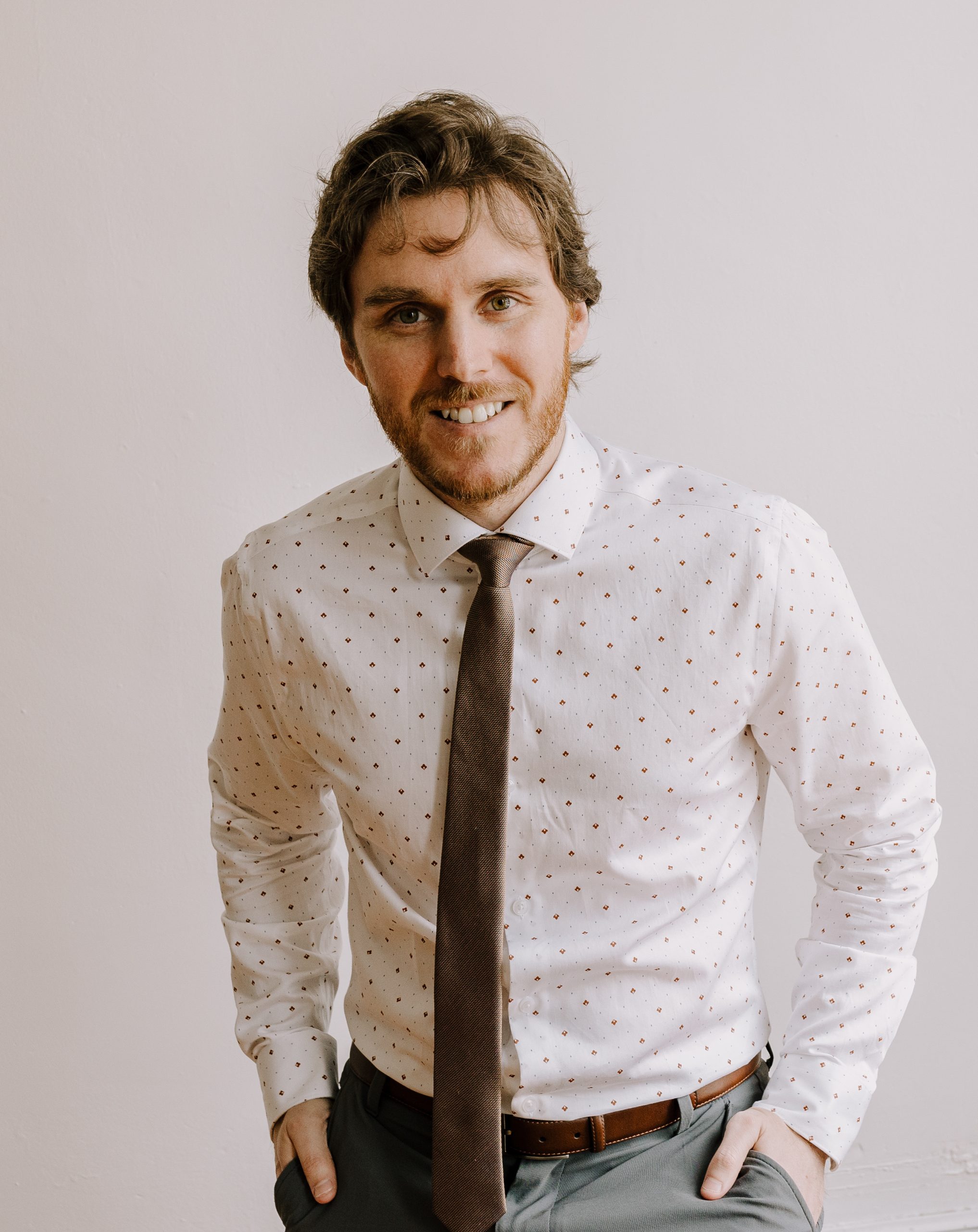 Connor is wearing a light polka dotted shirt with brown tie, belt and grey pants. he has his hands in his pockets as he leans forward a little smiling.