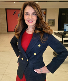 Leslie Sorensen Leslie wears a navy jacket with gold buttons over a red dress. She is smiling with her hands on her hips.